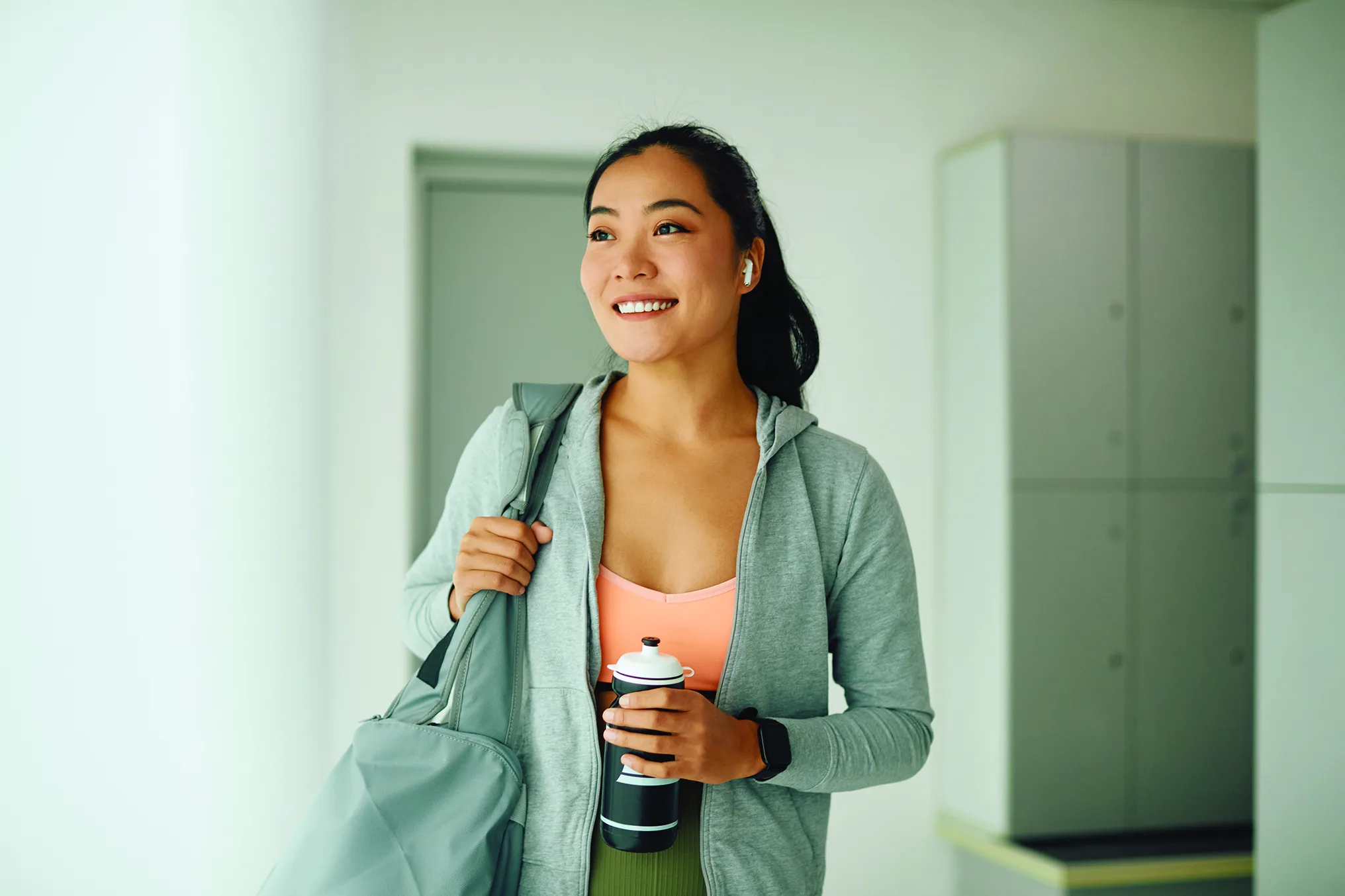 Woman smiling in an empty locker room, she is wearing an athletic top with a sweater unzipped where you can see her olive-green athletic leggings. She is wearing an apple watch and holding her water bottle with her left arm while her right arm braces her gym bag on her right shoulder. She isn't looking at the camera but possible out a window.