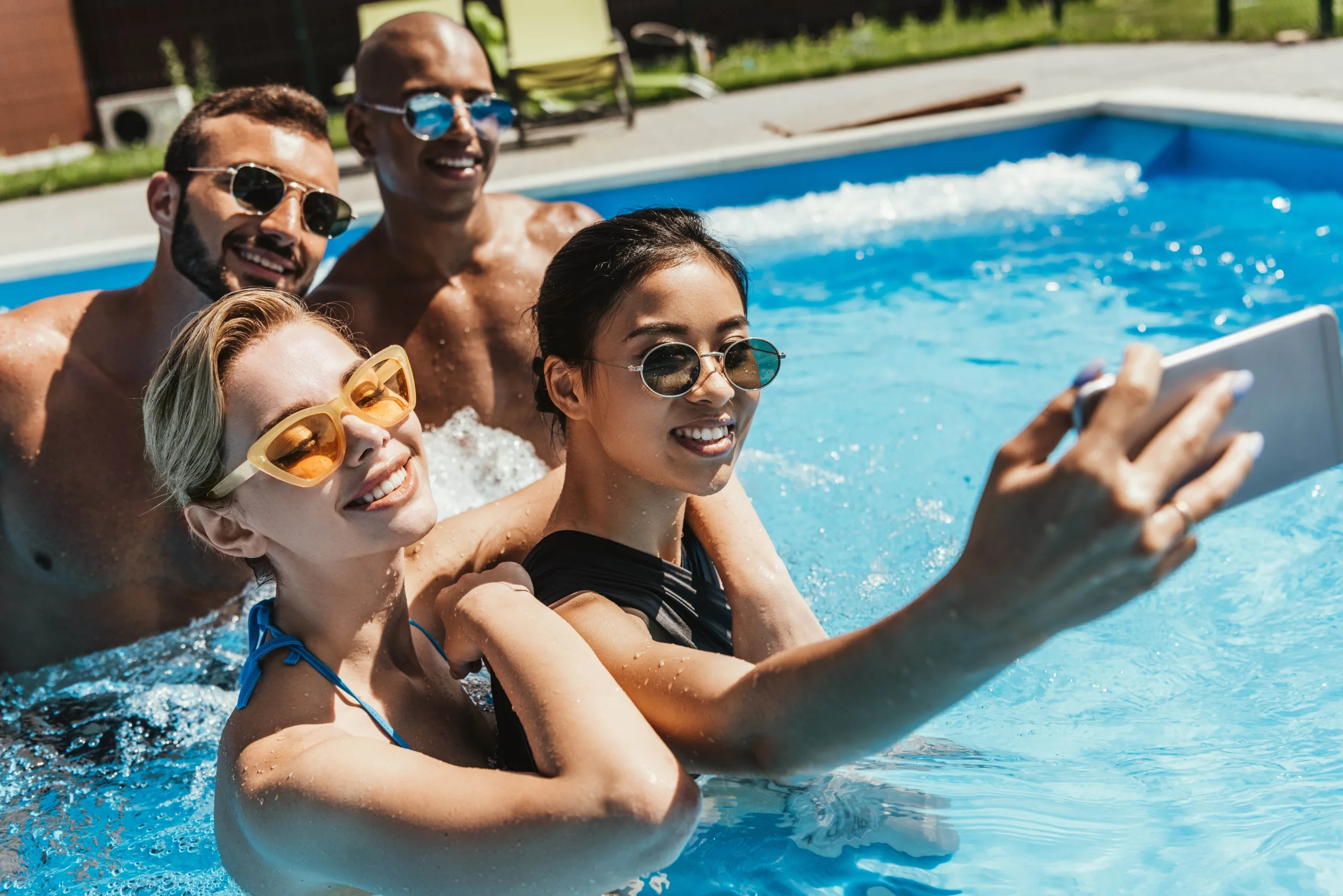 4 friends in pool smiling taking a selfie; two men and two women all with sunglasses on and some candid splashes.