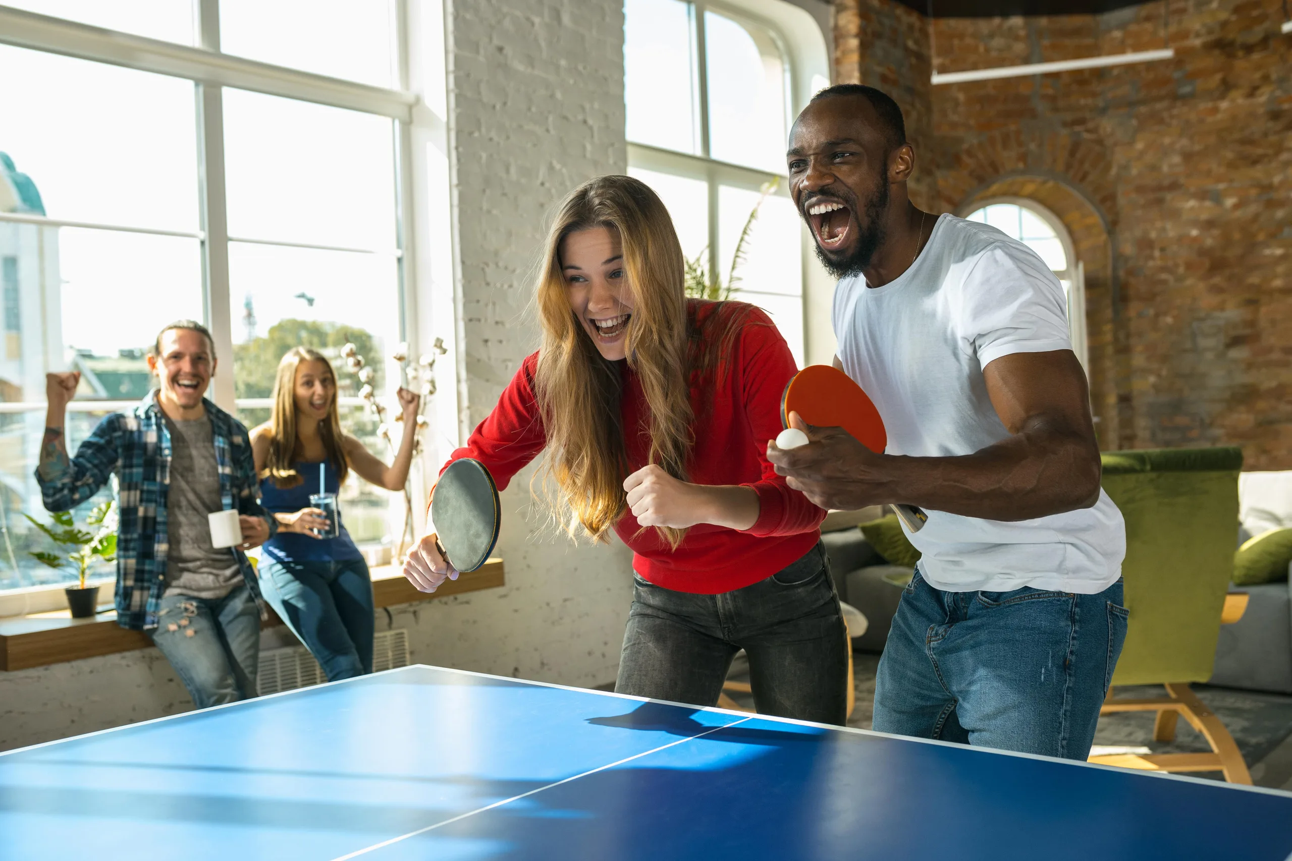 Man and woman playing table tennis in the clubhouse. Their friends are in the background cheering them on holding refreshments and leaning on a window seal with a city in the backdrop.