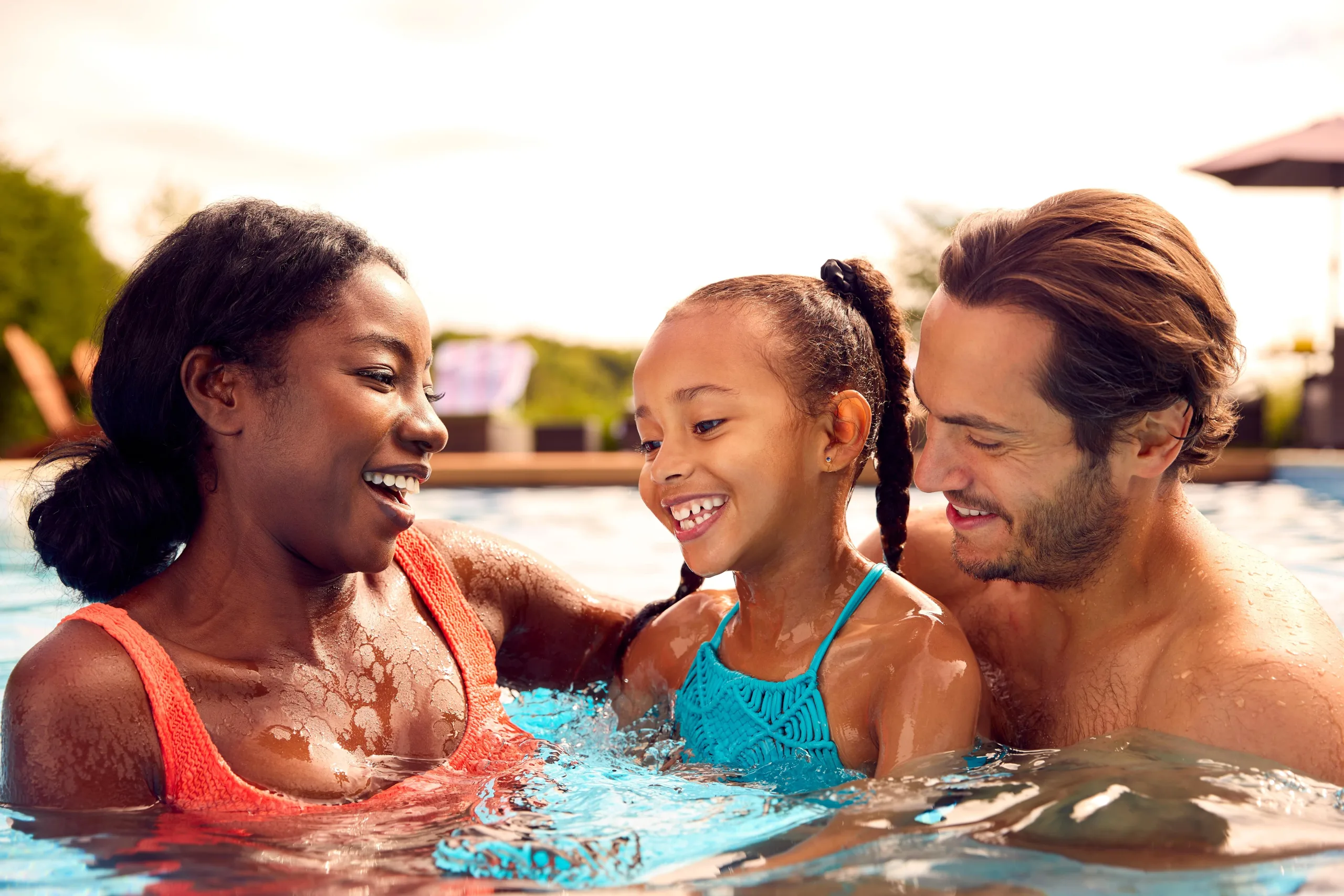 Family photo in the pool candid moment. Dark-skinned woman in a coral bikini on the left with thick curly hair. She is gazing at her daughter who has lighter skinned two braided pigtails in a bright blue swimsuit being held by her dad a fair-skinned man with a stubble beard and longer hair staring and smiling at his daughter.